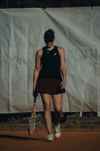 Athletic woman walking on clay court with tennis racket. Dark sportswear and visor create a fashionable look.