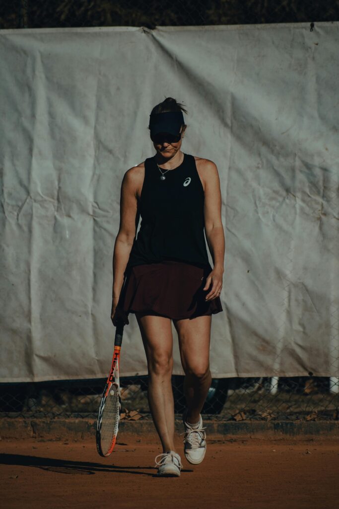 Athletic woman walking on clay court with tennis racket. Dark sportswear and visor create a fashionable look.